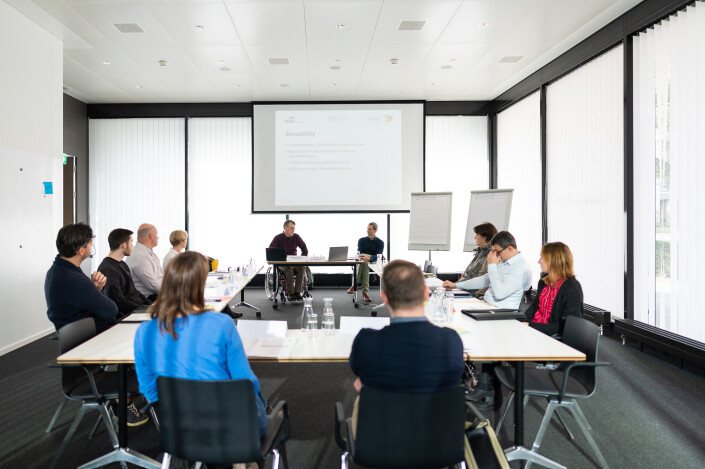 Herbert Bichsel und Pascal Güntensperger stimmen die Teilnehmenden auf das Praxisseminar ein. Die Teilnehmenden sitzen an den Tischen, die sich in einer U-Form befinden.