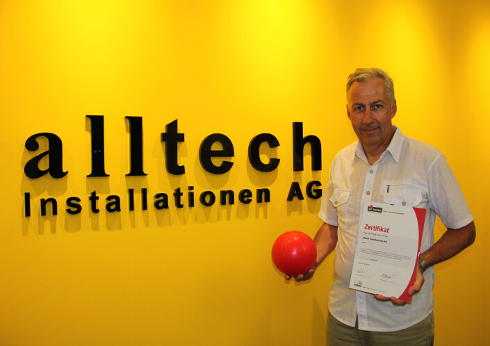 Roland Ming, Mitglied der Geschäftsleitung alltech Installationenen AG mit iPunkt-Zertifikat und rotem Ball vor Firmenlogo auf gelbem Hintergrund.